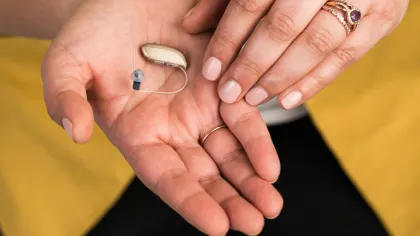 Person holding small beige hearing aid device in one hand with the other hand touching it, wearing a yellow jacket.
