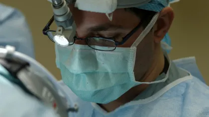 Surgeon wearing surgical mask, glasses, and headlight focusing on a medical procedure in operating room.