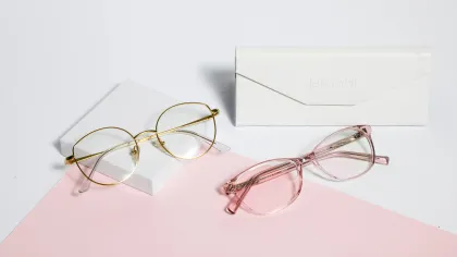 Two pairs of eyeglasses, one gold metal frame and one pink translucent frame, with a white lensabl case on white and pink background.