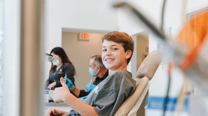 Smiling boy with braces giving thumbs up while sitting in a dental chair at clinic with staff in background.
