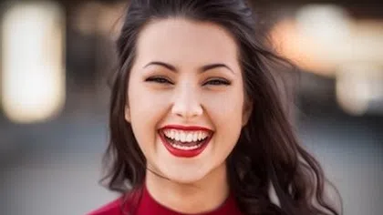 Portrait of a young woman with dark hair smiling brightly, wearing red lipstick and a red top outdoors.
