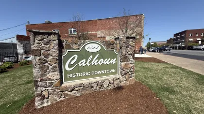 Stone sign reading Calhoun Historic Downtown 1852 with brick buildings and clear blue sky in background.