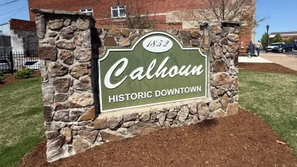 Welcome sign for Calhoun Historic Downtown, featuring stone design and green background under blue sky.