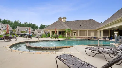 Residential community pool area with lounge chairs, umbrellas, and houses in the background under clear sky.