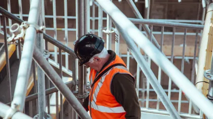 Construction worker wearing an orange safety vest and black helmet inspecting scaffolding at site.