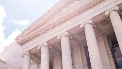 Classical building facade with tall marble columns and an eagle emblem in sunlight against a blue sky.