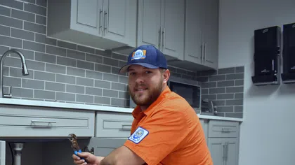 Estes Plumber in orange uniform kneeling by kitchen sink holding a pipe wrench in a modern grey and white kitchen.