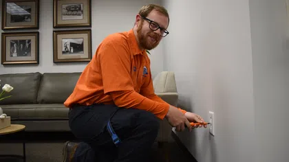 Estes Services Electrician in orange shirt working on wall outlet in modern living room with couch and framed photos.