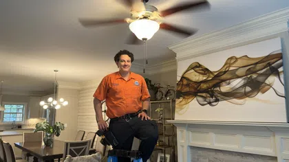 Handyman in orange shirt standing on ladder in living room with modern decor and ceiling fan spinning above.
