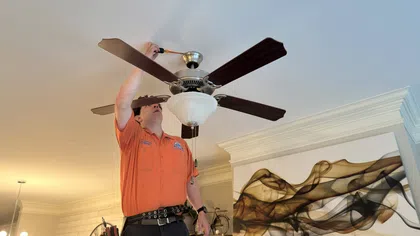 Estes Services Technician in orange shirt installing a ceiling fan light fixture in a modern living room with abstract wall art.