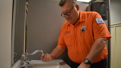 Estes Plumber worker in an orange shirt inspecting a faucet in a public restroom with gray partitions.