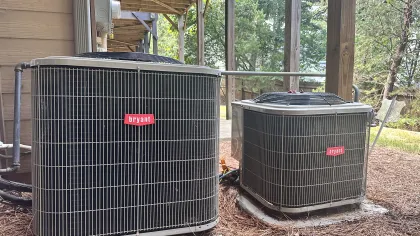 Two Bryant brand outdoor HVAC air conditioning units installed beside a wooden house under a deck.