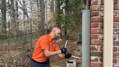 Estes Services Technician in orange shirt installing electrical wiring on brick house exterior in wooded backyard area.