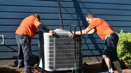 Two Estes Services Installers in orange shirts installing a Carrier air conditioning unit outside a blue house on a sunny day.
