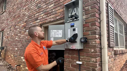Electrician in orange shirt inspecting an outdoor electrical panel mounted on a brick wall of a residential building.