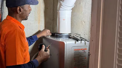 Technician in orange shirt inspecting and repairing a residential water heater with tools in a basement