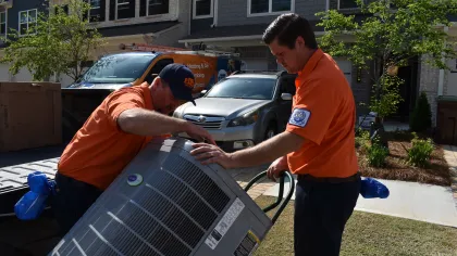 Two technicians in orange uniforms installing or repairing an outdoor air conditioning unit outside a residential home.