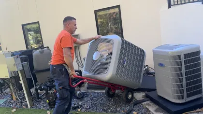 Estes Services Technician in orange uniform carrying a Carrier air conditioning unit on a red dolly near a building exterior.