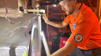 Estes Services Technician wearing orange uniform and headlamp inspects HVAC system in attic with tools and equipment nearby.