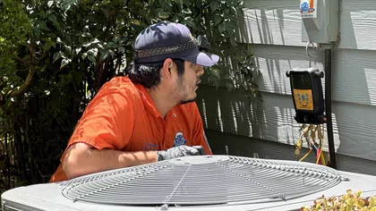 Estes Services Technician in orange uniform inspecting outdoor air conditioning unit beside a house wall with diagnostic equipment.