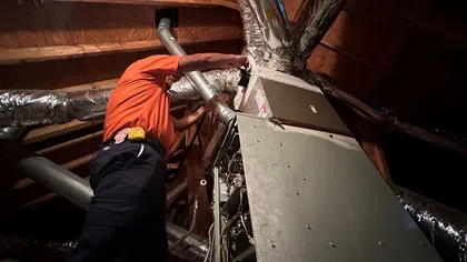 Estes Services Technician in orange shirt repairing HVAC furnace unit inside attic with exposed wooden beams and insulated ductwork.