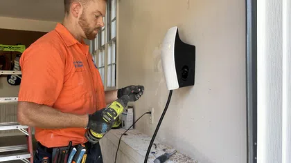 Technician in orange shirt installing wall-mounted electric vehicle charger using power drill and tools in garage.
