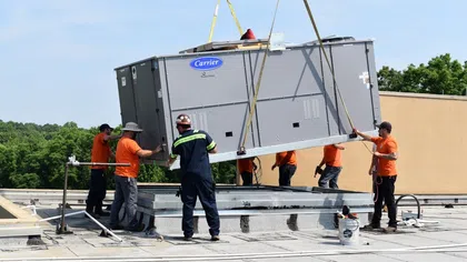 Workers installing a large Carrier HVAC unit on a rooftop using crane and safety gear on a sunny day.