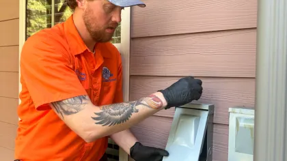 Estes Services Air Repair Technician in orange uniform and black gloves inspecting an outdoor electrical outlet on a beige siding wall.