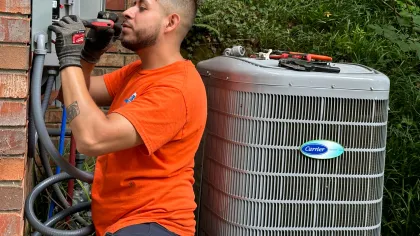 Estes HVAC Technician in orange installing an outdoor Carrier air conditioning unit by brick wall
