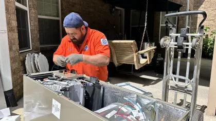 Technician in orange shirt repairing HVAC unit outdoors with tools and equipment under covered patio.
