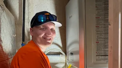 Technician in an orange shirt repairs plumbing with pipes and a white bucket in a utility room with reflective insulation.