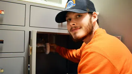 Estes Services plumber in orange sweatshirt and cap fixing pipe under sink cabinet indoors looking at camera.