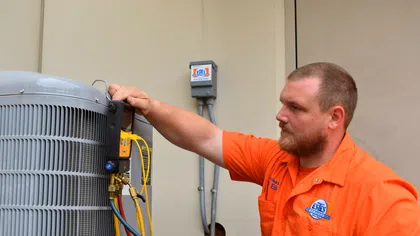 Technician in orange uniform inspecting and measuring air conditioning unit with gauges and hoses.