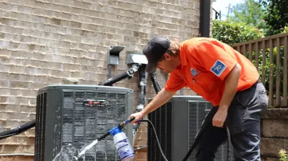 Estes Services Technician in orange shirt cleaning an outdoor air conditioning unit with a spray bottle and hose against a brick wall.