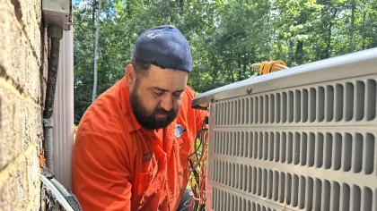 Estes Heating and cooling technician in orange shirt repairing an outdoor air conditioning unit on a sunny day