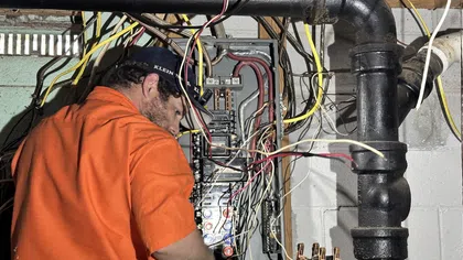 Estes Services Electrician working on circuit breaker panel with numerous wires in a basement setting.