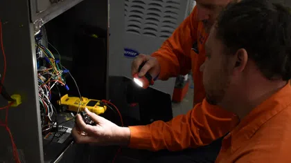 Two technicians in orange shirts repairing an HVAC unit using a flashlight and multimeter on exposed wiring.