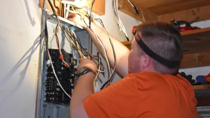 Estes Electrician in an orange shirt working on electrical panel wiring with exposed wires and tools in a wooden workshop.