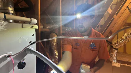 Estes Services Technician wearing headlamp inspecting HVAC system inside attic with exposed insulation and wooden beams.