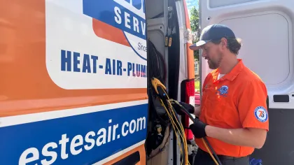 Estes Services Technician in orange uniform working with tools inside Estes Air service van labeled Heat-Air-Plumbing and estesair.com.