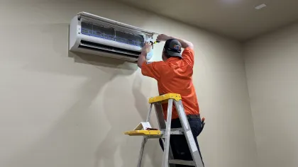 Technician in orange shirt servicing a wall-mounted air conditioner while standing on a ladder indoors.