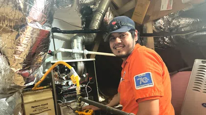 Estes Services Technician in orange shirt servicing HVAC furnace with diagnostic tools in attic space, smiling at camera.