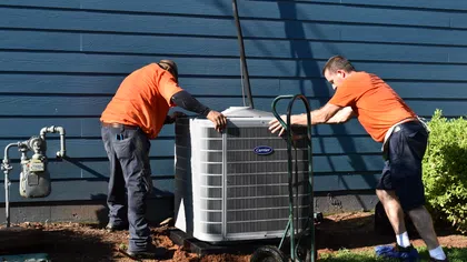 Two Estes Services Technicians in orange shirts installing a Carrier air conditioning unit outside a blue house on a sunny day.