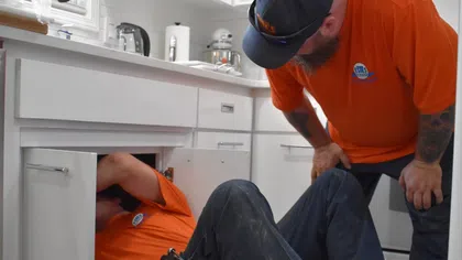 Two Estes Services plumbers in orange shirts working under a kitchen sink with tools and equipment visible.