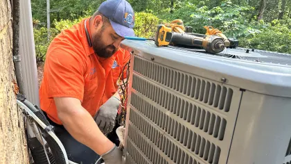 Estes Services Technician in orange shirt repairing an outdoor air conditioning unit with tools on top near a wooded area.