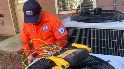 HVAC technician in an orange uniform performing AC maintenance on an outdoor air conditioning unit using gauges and diagnostic tools beside a home.