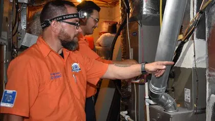 Two HVAC technicians in orange shirts inspecting and pointing at a furnace system in a utility room.