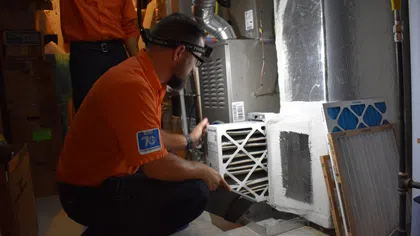 HVAC technician in orange shirt replacing furnace air filter in a residential basement setting.