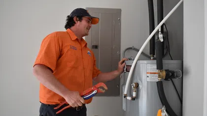 Estes Services Plumber in orange uniform holding wrench inspecting a gray water heater in a home utility room.