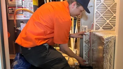 Estes Services Technician in orange shirt and cap inspecting and repairing HVAC system in utility closet.